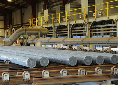 The image shows an industrial setting with multiple large aluminum billets lined up on a conveyor system ready for processing or transport.