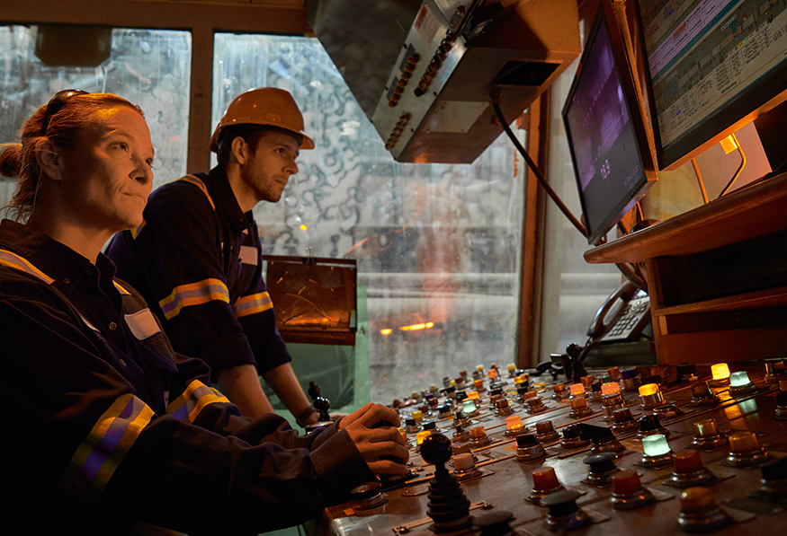 Two people wearing safety helmets and reflective work gear operate a control panel with numerous buttons and switches, focused intently on their task.