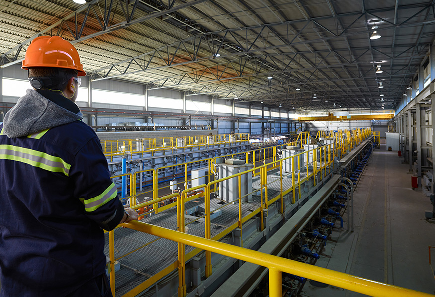 A person wearing a hard hat and safety vest observes operations in a large industrial factory with machinery, safety railings, and bright overhead lighting.