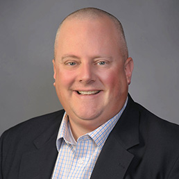 A smiling person with a bald head, wearing a business suit with a checkered shirt and tie, poses for a professional headshot against a gray background.