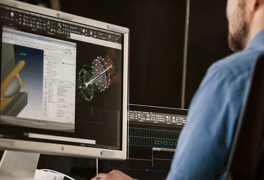 A person in a blue shirt is working on a complex CAD model displayed on dual computer monitors, likely engaged in engineering or design work.