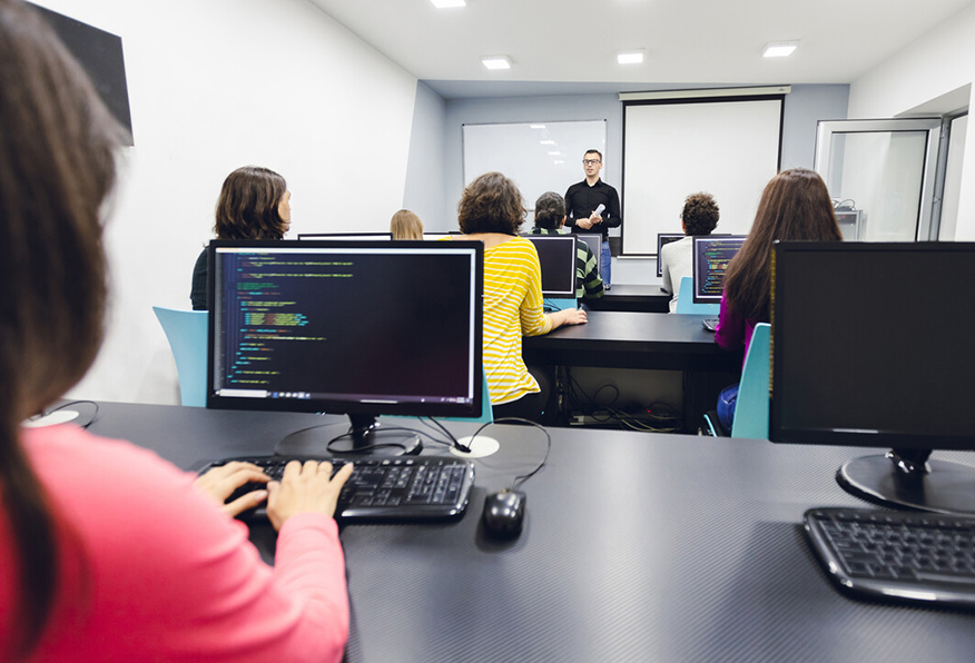 A person teaches computer programming to students in a modern classroom with monitors displaying code. The students focus on the lesson, some taking notes.