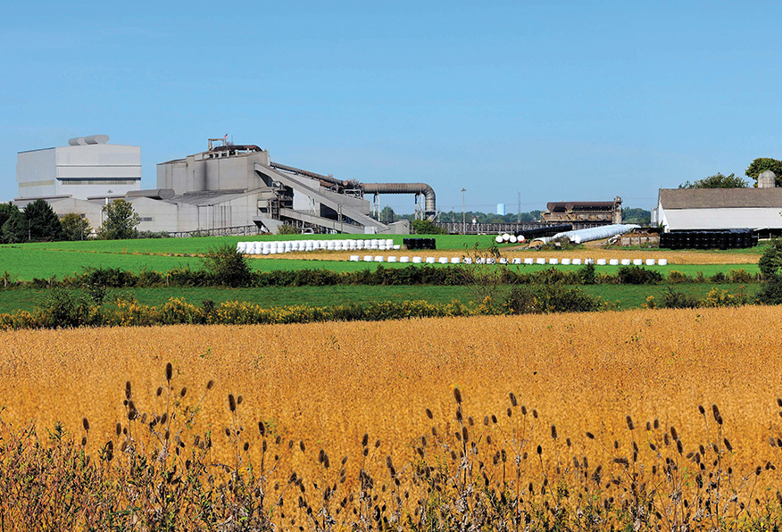 A large industrial facility with cylindrical storage tanks and conveyor belts stands behind a golden field under a clear blue sky. No people are visible.