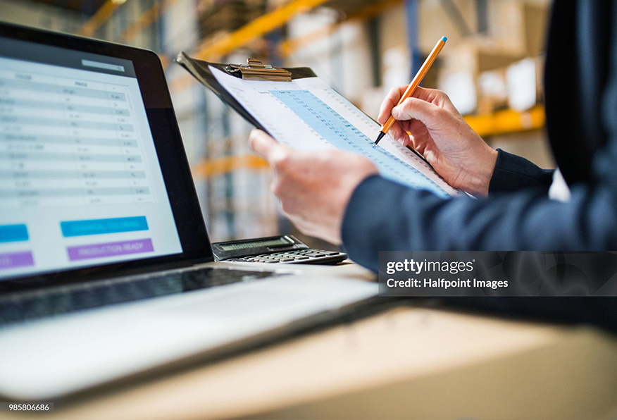 A person is checking a document on a clipboard while a laptop with charts on the screen sits nearby, possibly in a warehouse environment.