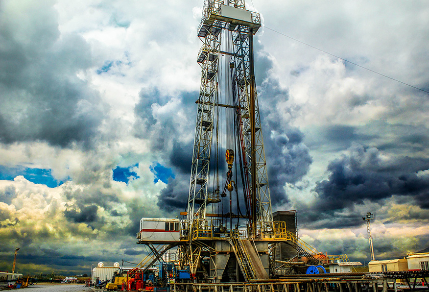 An oil drilling rig under a dramatic cloudy sky. Industrial equipment and structures are visible with some workers wearing safety gear on the site.