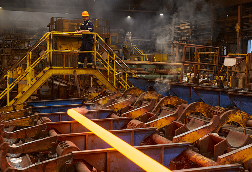 A person in safety gear oversees molten material in a steel mill. Industrial equipment surrounds the area with steam and a glowing orange metal piece prominent.