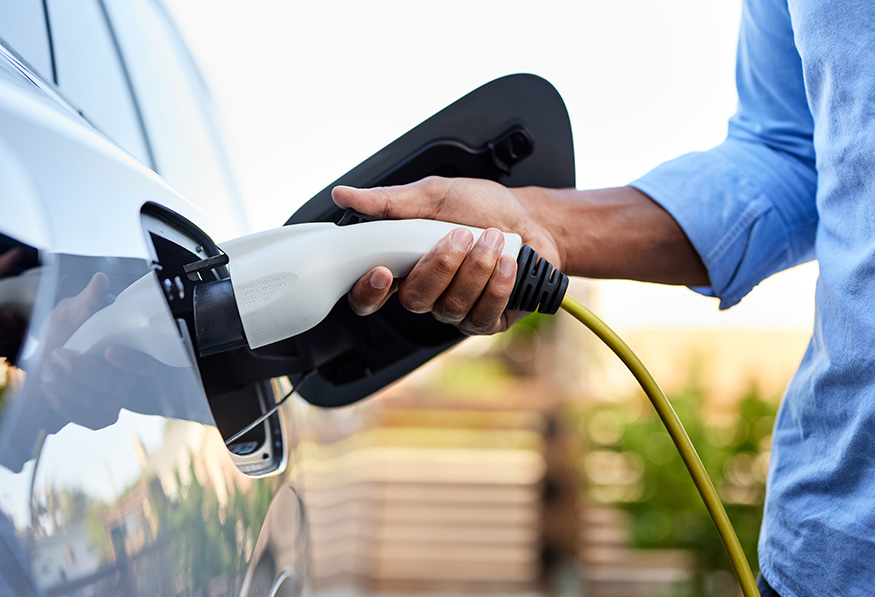 A person is plugging a charging connector into the port of a white electric vehicle against a blurred outdoor background, indicating green transportation.