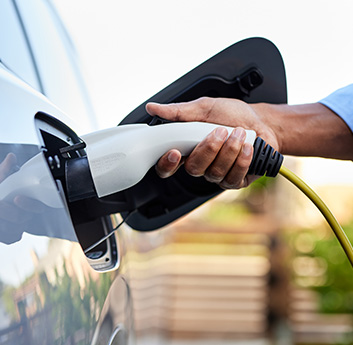 A person is plugging an electric vehicle (EV) charging cable into a car's charging port. Focus on hands, cable, and charger under natural lighting.