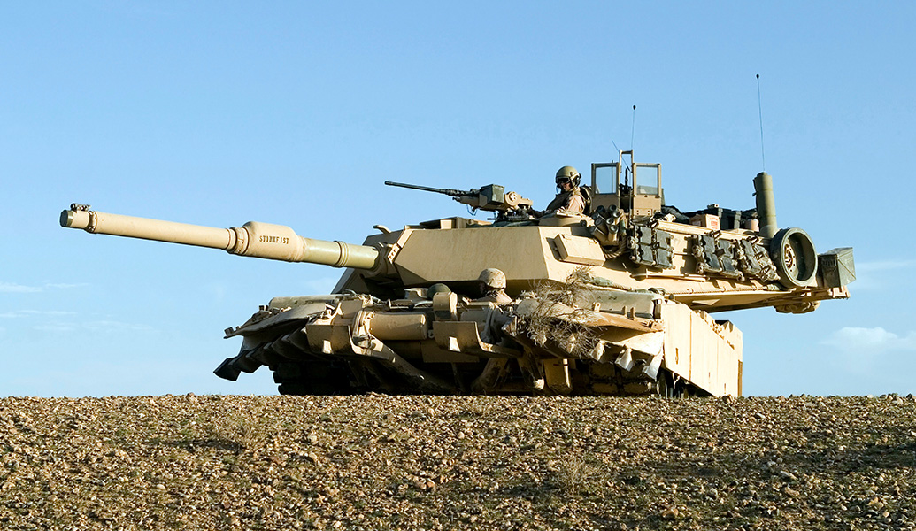 A military tank positioned on a rise with its cannon facing forward. One person is visible in tactical gear atop the tank, under a clear sky.