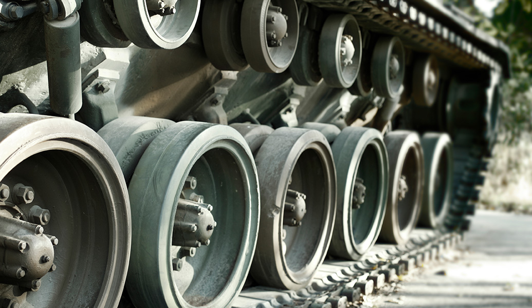 Close-up of a military tank's caterpillar tracks and road wheels, showing detailed tread and suspension components, with a blurred natural background.