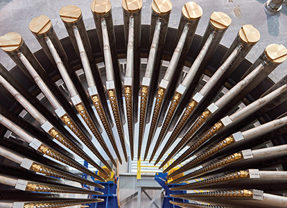 This image shows a collection of long, cylindrical metal rods arranged in a radial pattern on a blue rack, with their threaded ends capped in brass.