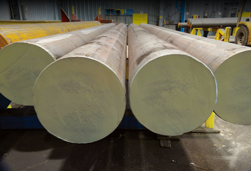 Large industrial steel pipes rest on blue supports inside a warehouse with a yellow floor marking and additional machinery in the background.