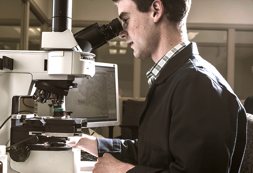 A person is using a sophisticated microscope in a laboratory setting, with a computer monitor in the background, indicating a research or scientific analysis.